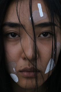 A close-up of a person's face featuring strands of dark hair partially obscuring their eyes and forehead. The skin has patches of white and beige makeup or paint applied in various shapes on the forehead and cheeks. The expression is neutral and introspective, and the lighting is dim, creating a moody atmosphere.