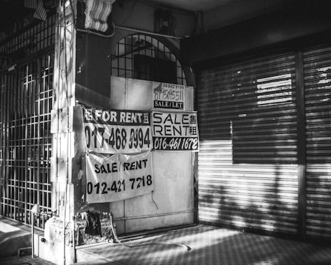 A black and white image of a closed storefront with metal shutters. Several signs with 'For Rent' and 'Sale/Rent' are prominently displayed, including phone numbers for contact. The exterior appears worn, with shadows cast across the scene.