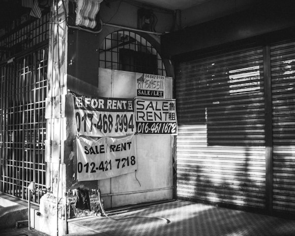 A black and white image of a closed storefront with metal shutters. Several signs with 'For Rent' and 'Sale/Rent' are prominently displayed, including phone numbers for contact. The exterior appears worn, with shadows cast across the scene.