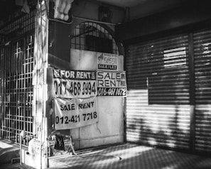 A black and white image of a closed storefront with metal shutters. Several signs with 'For Rent' and 'Sale/Rent' are prominently displayed, including phone numbers for contact. The exterior appears worn, with shadows cast across the scene.