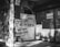 A black and white image of a closed storefront with metal shutters. Several signs with 'For Rent' and 'Sale/Rent' are prominently displayed, including phone numbers for contact. The exterior appears worn, with shadows cast across the scene.