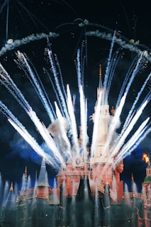 Vibrant fireworks explode in the night sky above a large, illuminated castle, creating a celebratory and magical atmosphere.