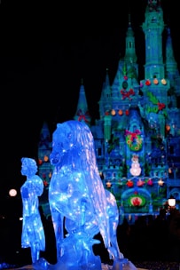 An illuminated ice sculpture of a figure standing next to a horse is displayed in front of a colorfully lit castle at night. The castle is decorated with festive ornaments, creating a magical and enchanting atmosphere.