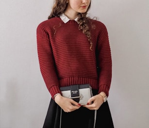 A person with long curly hair is wearing a red knitted sweater over a white collared shirt. They are holding a small Prada handbag. The person's outfit includes a black skirt, and they are wearing a watch on their left wrist.