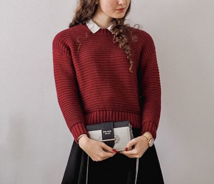 A person with long curly hair is wearing a red knitted sweater over a white collared shirt. They are holding a small Prada handbag. The person's outfit includes a black skirt, and they are wearing a watch on their left wrist.