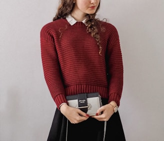 A person with long curly hair is wearing a red knitted sweater over a white collared shirt. They are holding a small Prada handbag. The person's outfit includes a black skirt, and they are wearing a watch on their left wrist.