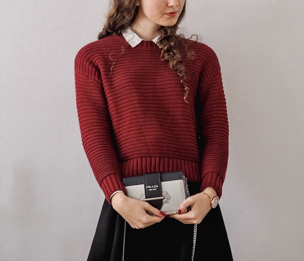 A person with long curly hair is wearing a red knitted sweater over a white collared shirt. They are holding a small Prada handbag. The person's outfit includes a black skirt, and they are wearing a watch on their left wrist.