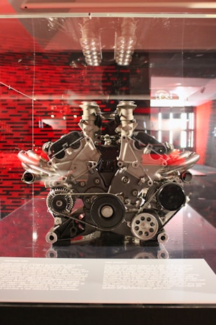 A high-performance engine is displayed on a platform, showcasing its complex mechanical components. The engine appears to be a V12 configuration with a mix of metal parts, belts, and pulleys. The backdrop features a red and black pattern, contributing to a modern and technical ambiance.