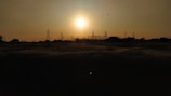 The setting sun casts a warm glow over a landscape dotted with electrical towers and power lines. The silhouettes of the towers create a striking contrast against the bright sky, while a few birds can be seen flying near the horizon.