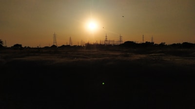 The setting sun casts a warm glow over a landscape dotted with electrical towers and power lines. The silhouettes of the towers create a striking contrast against the bright sky, while a few birds can be seen flying near the horizon.