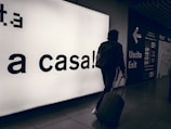 A person wearing a hooded jacket and carrying a backpack pulls a suitcase through an airport terminal. They walk past a large illuminated sign with the words 'a casa!' alongside directional signs in both Italian and English. The scene is dimly lit, emphasizing the glow of the signage.