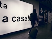 A person wearing a hooded jacket and carrying a backpack pulls a suitcase through an airport terminal. They walk past a large illuminated sign with the words 'a casa!' alongside directional signs in both Italian and English. The scene is dimly lit, emphasizing the glow of the signage.