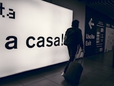 A person wearing a hooded jacket and carrying a backpack pulls a suitcase through an airport terminal. They walk past a large illuminated sign with the words 'a casa!' alongside directional signs in both Italian and English. The scene is dimly lit, emphasizing the glow of the signage.