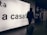 A person wearing a hooded jacket and carrying a backpack pulls a suitcase through an airport terminal. They walk past a large illuminated sign with the words 'a casa!' alongside directional signs in both Italian and English. The scene is dimly lit, emphasizing the glow of the signage.