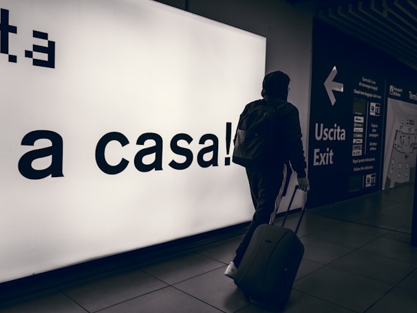 A person wearing a hooded jacket and carrying a backpack pulls a suitcase through an airport terminal. They walk past a large illuminated sign with the words 'a casa!' alongside directional signs in both Italian and English. The scene is dimly lit, emphasizing the glow of the signage.