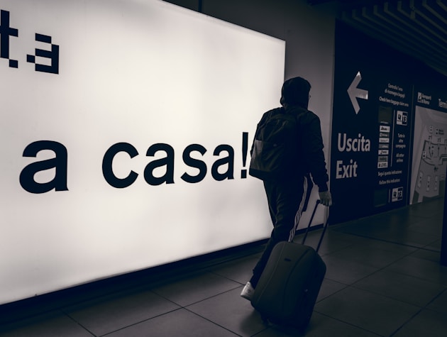 A person wearing a hooded jacket and carrying a backpack pulls a suitcase through an airport terminal. They walk past a large illuminated sign with the words 'a casa!' alongside directional signs in both Italian and English. The scene is dimly lit, emphasizing the glow of the signage.