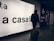 A person wearing a hooded jacket and carrying a backpack pulls a suitcase through an airport terminal. They walk past a large illuminated sign with the words 'a casa!' alongside directional signs in both Italian and English. The scene is dimly lit, emphasizing the glow of the signage.