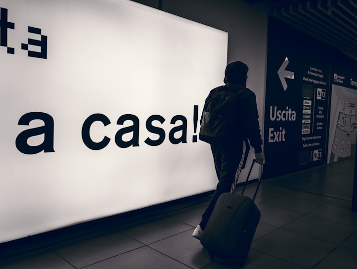 A person wearing a hooded jacket and carrying a backpack pulls a suitcase through an airport terminal. They walk past a large illuminated sign with the words 'a casa!' alongside directional signs in both Italian and English. The scene is dimly lit, emphasizing the glow of the signage.