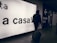 A person wearing a hooded jacket and carrying a backpack pulls a suitcase through an airport terminal. They walk past a large illuminated sign with the words 'a casa!' alongside directional signs in both Italian and English. The scene is dimly lit, emphasizing the glow of the signage.