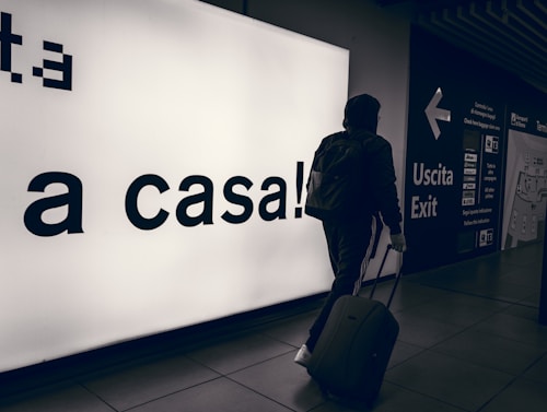 A person wearing a hooded jacket and carrying a backpack pulls a suitcase through an airport terminal. They walk past a large illuminated sign with the words 'a casa!' alongside directional signs in both Italian and English. The scene is dimly lit, emphasizing the glow of the signage.