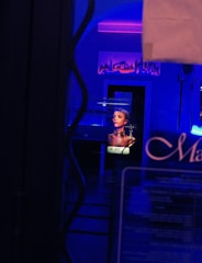 A dimly lit interior with neon lights casting a blue and purple hue. A reflective surface shows part of a perfume advertisement featuring a person, with the word 'Dior' visible. The setting is somewhat modern, with an urban city skyline artwork on the wall.