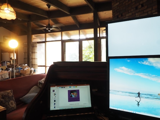 A cozy living room with a wooden ceiling and beams, featuring a large window with sheer curtains that allow natural light to fill the space. There are two computer monitors on a wooden desk, one displaying a bright image of a person running on a beach and the other showing a presentation slide. A lamp with a warm glow hangs from the ceiling, adding to the inviting atmosphere. A sofa with floral cushions is visible, along with baskets and various household items on a table.