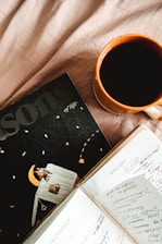 A cup of coffee in an orange mug sits on a soft, beige fabric surface next to an open planner and a magazine titled 'Lessons of life.' The planner displays handwritten notes for December and January, with colorful highlights and checkmarks. The magazine cover features a whimsical illustration of a person sitting on a moon.