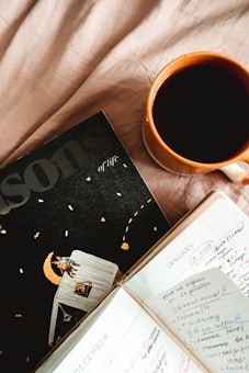 A cup of coffee in an orange mug sits on a soft, beige fabric surface next to an open planner and a magazine titled 'Lessons of life.' The planner displays handwritten notes for December and January, with colorful highlights and checkmarks. The magazine cover features a whimsical illustration of a person sitting on a moon.