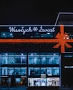A building is decorated with festive lights, including a large orange bow and the words 'Wesołych Świąt' in white lights. The facade has a modern design with large glass windows. Several stores, including a Domino's Pizza, are visible on the ground floor. The building has a dark exterior contrasted with bright interior lighting.
