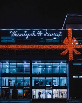A building is decorated with festive lights, including a large orange bow and the words 'Wesołych Świąt' in white lights. The facade has a modern design with large glass windows. Several stores, including a Domino's Pizza, are visible on the ground floor. The building has a dark exterior contrasted with bright interior lighting.