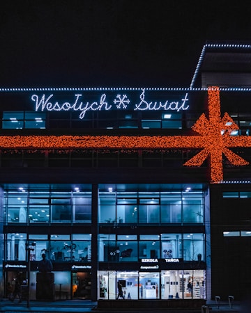 A building is decorated with festive lights, including a large orange bow and the words 'Wesołych Świąt' in white lights. The facade has a modern design with large glass windows. Several stores, including a Domino's Pizza, are visible on the ground floor. The building has a dark exterior contrasted with bright interior lighting.