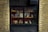 A brick wall contains a large black-framed window displaying various packaged goods on wooden shelves. The items include bags of pasta and boxes, arranged neatly against a red backdrop. The window reflects small lights or chandeliers from the ceiling.