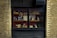 A brick wall contains a large black-framed window displaying various packaged goods on wooden shelves. The items include bags of pasta and boxes, arranged neatly against a red backdrop. The window reflects small lights or chandeliers from the ceiling.