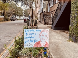 A residential street scene shows a sign in a garden bed near the sidewalk. The sign features a hand-drawn portrait of a smiling person and includes a quote in blue text: 'I've always wanted to have a neighbor just like you.' attributed to Fred Rogers. The background includes parked cars and houses with trees lining the street.