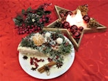 A festive table setting features a plate with an ornate holiday cake decorated with sprigs of pine, orange slices, star anise, and cinnamon sticks on a rich red tablecloth with a subtle floral pattern. Next to the cake, a wooden star-shaped candle holder is adorned with red baubles and pine cones, emitting a warm glow. Decorative foliage with red berries complements the scene, creating a cozy, holiday atmosphere.