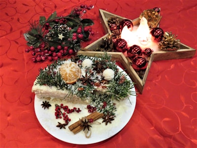 A festive table setting features a plate with an ornate holiday cake decorated with sprigs of pine, orange slices, star anise, and cinnamon sticks on a rich red tablecloth with a subtle floral pattern. Next to the cake, a wooden star-shaped candle holder is adorned with red baubles and pine cones, emitting a warm glow. Decorative foliage with red berries complements the scene, creating a cozy, holiday atmosphere.