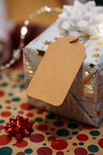 A gift with festive wrapping paper adorned with gold stars, a large white bow on top, and an attached blank brown tag. The background includes a surface covered with polka-dotted paper in red, green, and gold. A small red bow is placed nearby, contributing to the holiday theme.