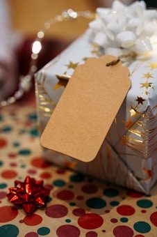 A gift with festive wrapping paper adorned with gold stars, a large white bow on top, and an attached blank brown tag. The background includes a surface covered with polka-dotted paper in red, green, and gold. A small red bow is placed nearby, contributing to the holiday theme.
