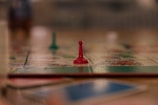 A close-up view of a board game with a focus on a single red pawn. The blurred background suggests other game pieces and a colorful game board. The scene appears to be on a table with a soft focus, giving a cozy and relaxed atmosphere.