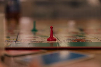 A close-up view of a board game with a focus on a single red pawn. The blurred background suggests other game pieces and a colorful game board. The scene appears to be on a table with a soft focus, giving a cozy and relaxed atmosphere.