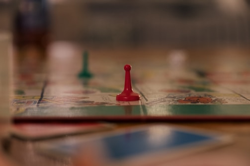 A close-up view of a board game with a focus on a single red pawn. The blurred background suggests other game pieces and a colorful game board. The scene appears to be on a table with a soft focus, giving a cozy and relaxed atmosphere.