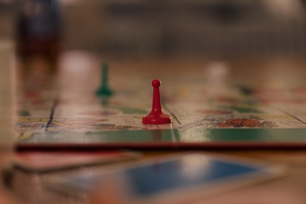 A close-up view of a board game with a focus on a single red pawn. The blurred background suggests other game pieces and a colorful game board. The scene appears to be on a table with a soft focus, giving a cozy and relaxed atmosphere.