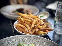 A plate filled with crispy, golden French fries next to a small dish of creamy dipping sauce. The background features other dishes blurred out, adding to a cozy, casual dining setting.