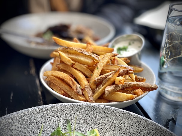 A plate filled with crispy, golden French fries next to a small dish of creamy dipping sauce. The background features other dishes blurred out, adding to a cozy, casual dining setting.