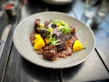 A gourmet dish is presented on a textured, circular plate. The dish features seared meat with a dark, charred exterior, accompanied by bright yellow mango chunks and garnished with fresh greens and herbs. Small legumes or grains surround the main components, providing contrast in color and texture. The setting is a polished dark wooden table with blurred elements in the background.