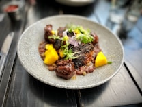 A gourmet dish is presented on a textured, circular plate. The dish features seared meat with a dark, charred exterior, accompanied by bright yellow mango chunks and garnished with fresh greens and herbs. Small legumes or grains surround the main components, providing contrast in color and texture. The setting is a polished dark wooden table with blurred elements in the background.