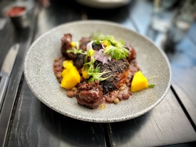 A gourmet dish is presented on a textured, circular plate. The dish features seared meat with a dark, charred exterior, accompanied by bright yellow mango chunks and garnished with fresh greens and herbs. Small legumes or grains surround the main components, providing contrast in color and texture. The setting is a polished dark wooden table with blurred elements in the background.
