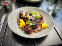 A gourmet dish is presented on a textured, circular plate. The dish features seared meat with a dark, charred exterior, accompanied by bright yellow mango chunks and garnished with fresh greens and herbs. Small legumes or grains surround the main components, providing contrast in color and texture. The setting is a polished dark wooden table with blurred elements in the background.