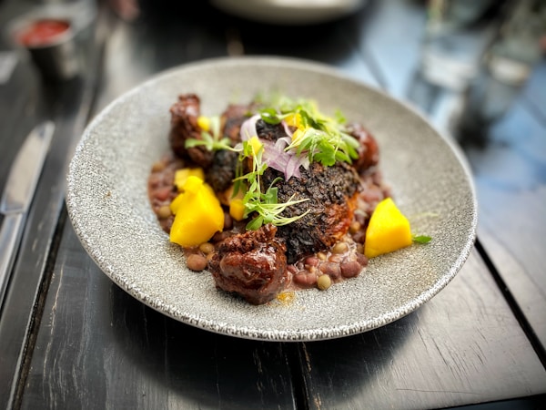 A gourmet dish is presented on a textured, circular plate. The dish features seared meat with a dark, charred exterior, accompanied by bright yellow mango chunks and garnished with fresh greens and herbs. Small legumes or grains surround the main components, providing contrast in color and texture. The setting is a polished dark wooden table with blurred elements in the background.