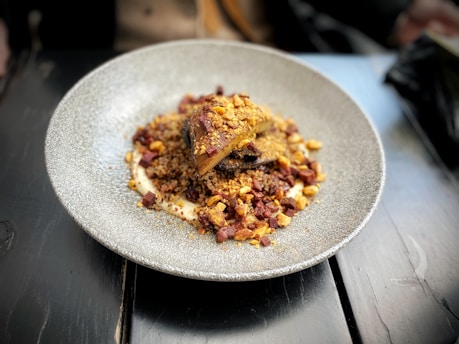 A plate with an elegantly arranged dish featuring what appears to be roasted meat or a similar protein, served over a bed of grains or couscous. The dish is garnished with diced pieces of vegetables or nuts, and is presented on a round, textured plate placed on a dark wooden table.